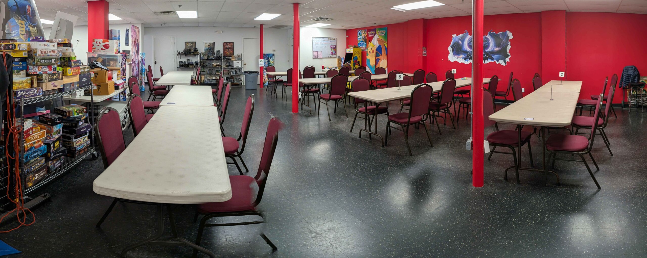 A brightly colored game room features rows of empty tables with chairs and a shelf stocked